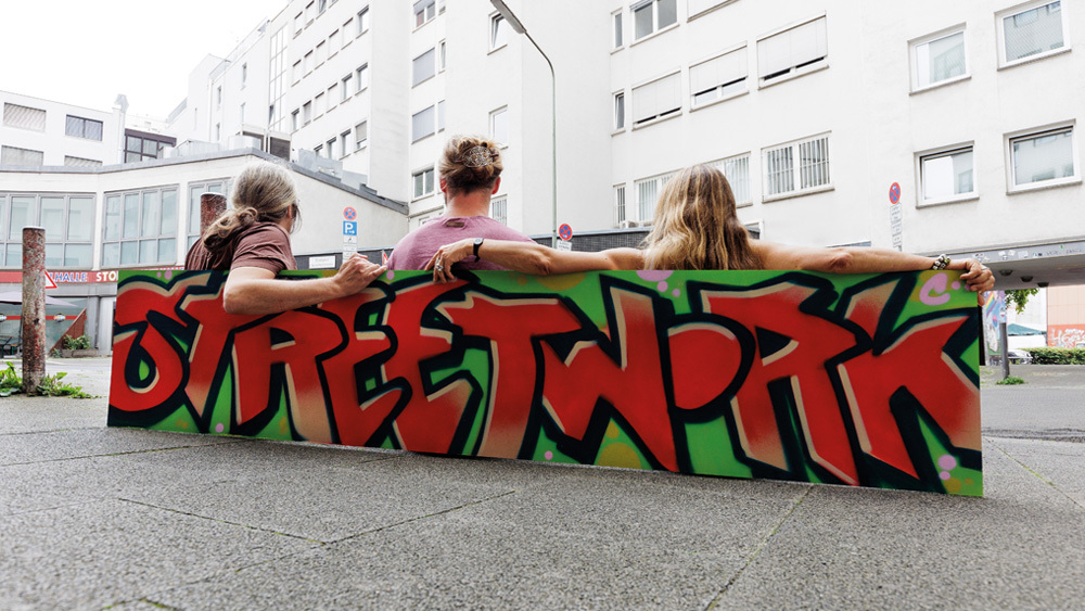 Drei Menschen sitzen vorm Beratungsladen auf der Straße hinter einem Schild, auf dem "Streetwork" steht.